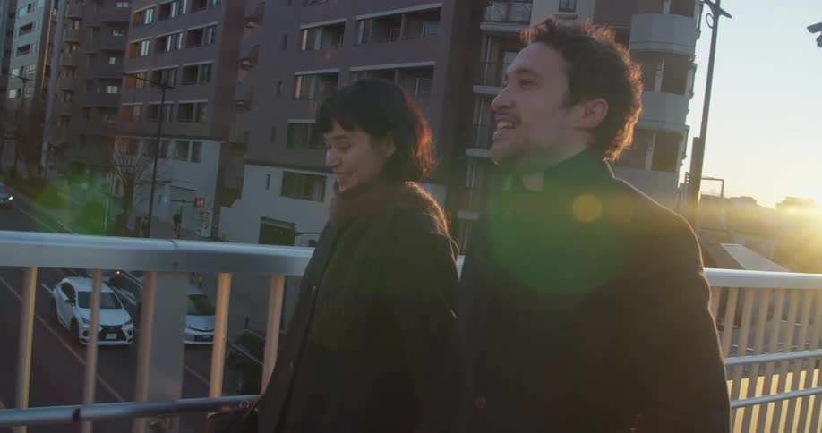 A man and a woman laugh and smile at each other while walking across a pedestrian bridge in a city