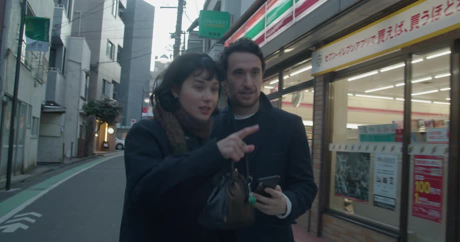 Male and female holding hands while walking down the street passing a convenience store and the male pulls his cell phone from his jacket pocket to look for directions