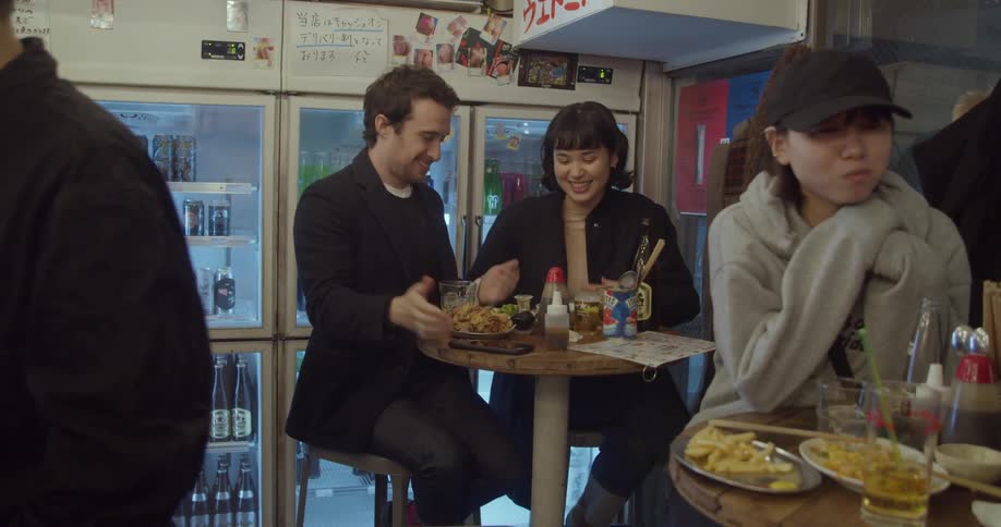 A couple sits in a restaurant and eats a meal as others wander around