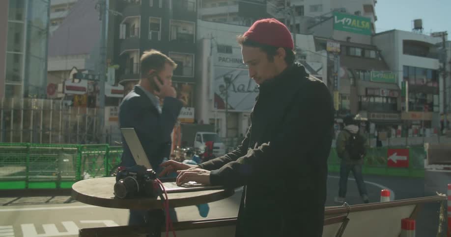 Man types on a laptop on an outdoor table as another man walks by talking on a cell phone