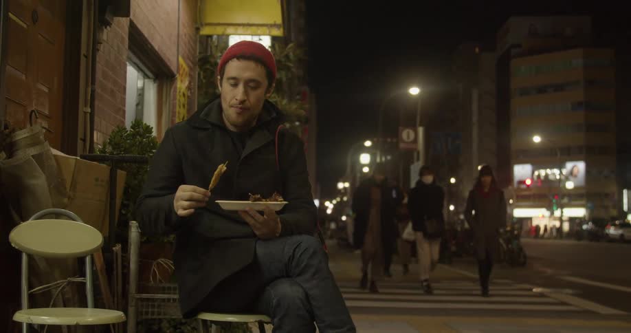 A man sits on a stool outside a restaurant holding a plate and eating food on a stick as people walk past