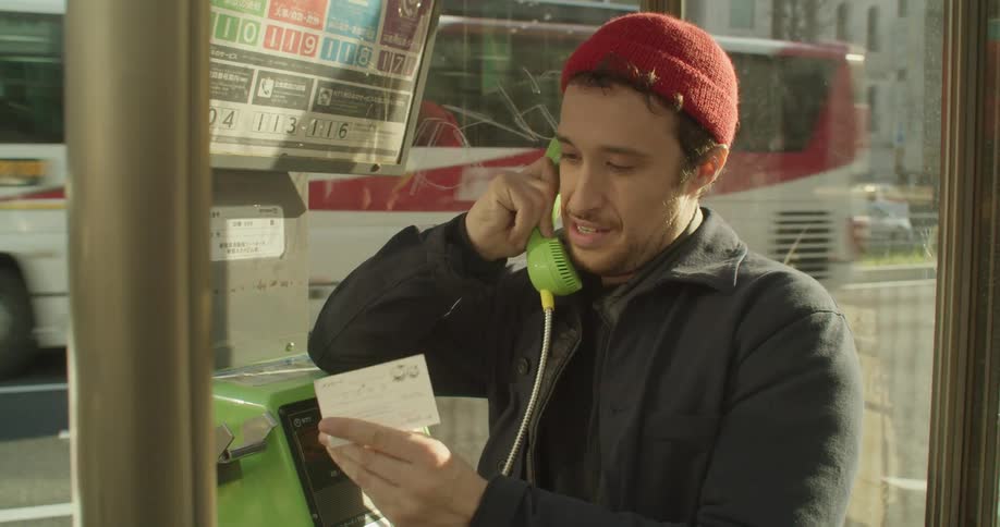 A man in a hat using a public payphone in an urban environment