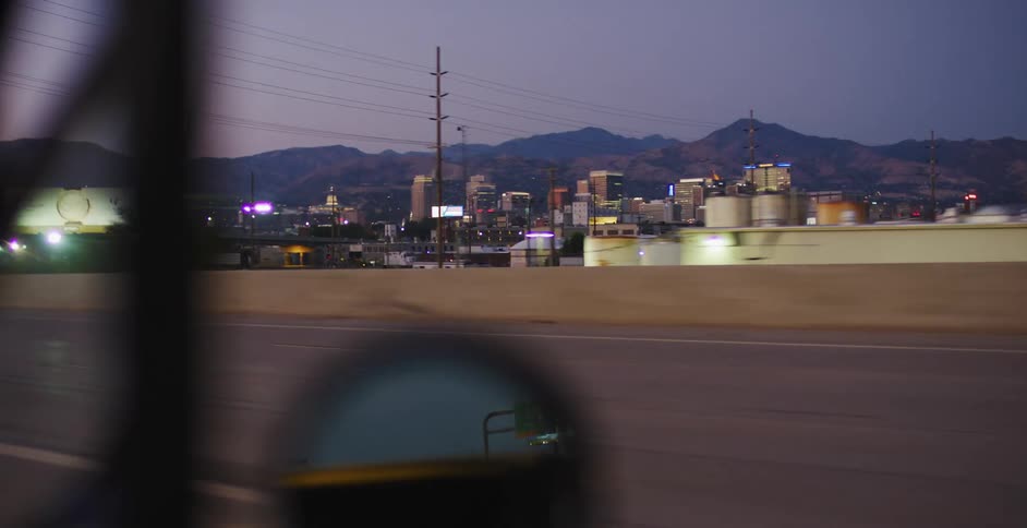 A vehicle drives down a highway with views of a nearby downtown and mountains