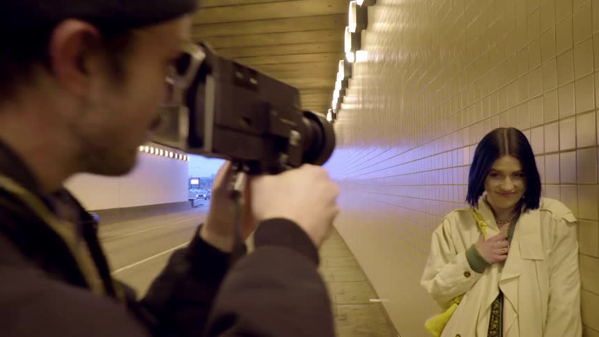 Man directs a woman to lean against a tunnel wall as he video records her