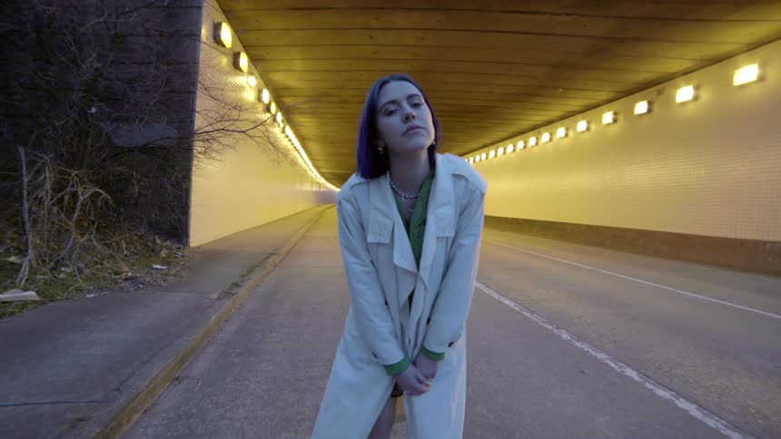 A white woman in an overcoat sways by the side of the road near a tunnel