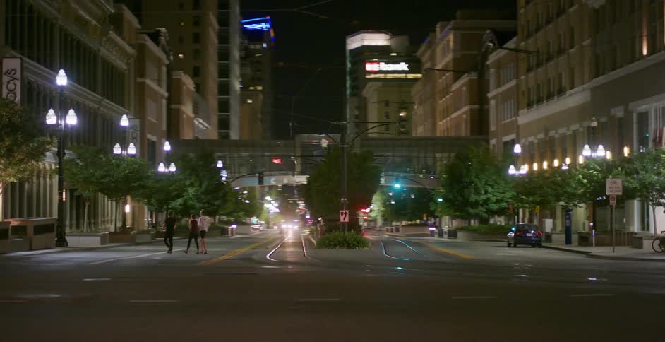 A group of three friends cross an empty city street lined with trees and lamp posts at night