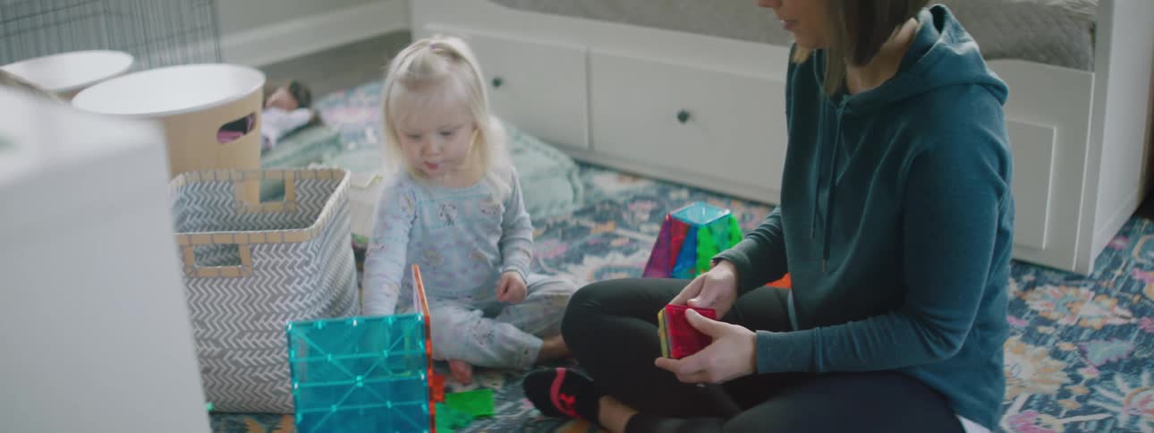Mother and two daughters sitting on the bedroom floor building with magnet tiles together as the toddler sucks on a lolly pop