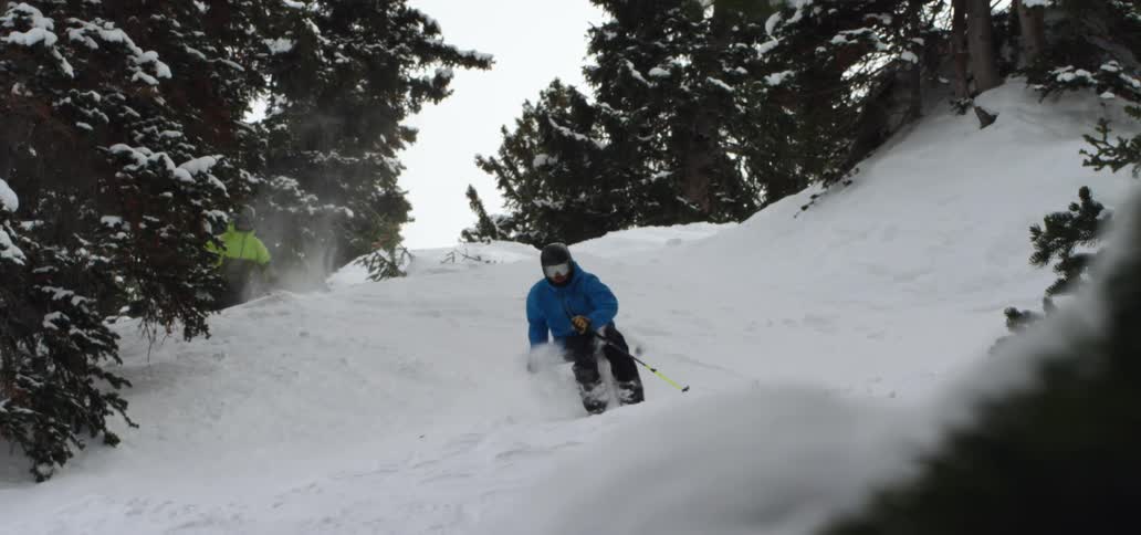 Two skiers jump as they come over the crest of a hill between some trees