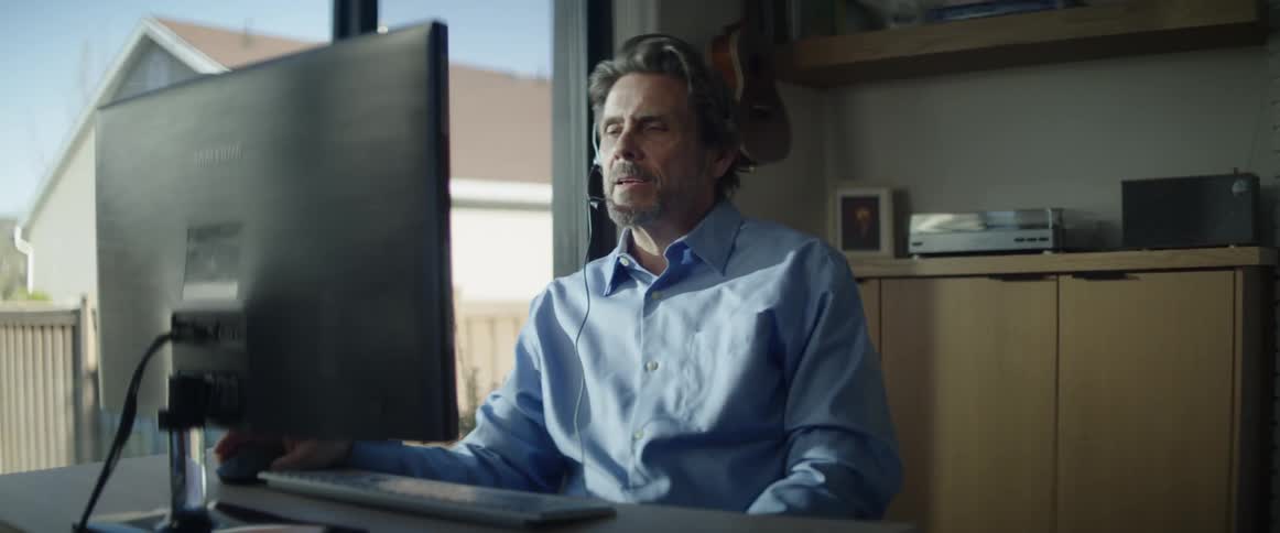A man sitting at his home office desk and computer puts on his headset and starts to talk