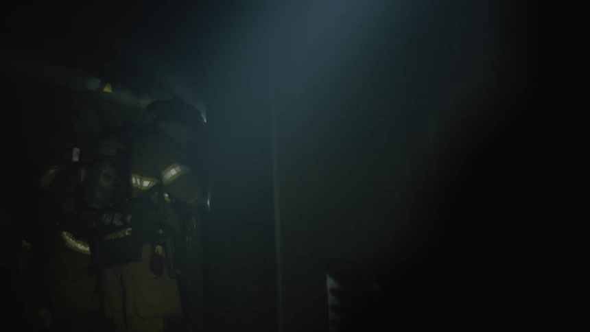 A firefighter in full gear uses a flashlight to search a dark room