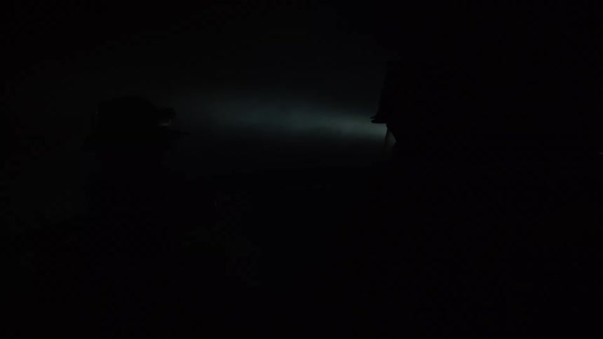 Two firefighters wearing gear stand inside a dark room as smoke wafts through a shaft of light