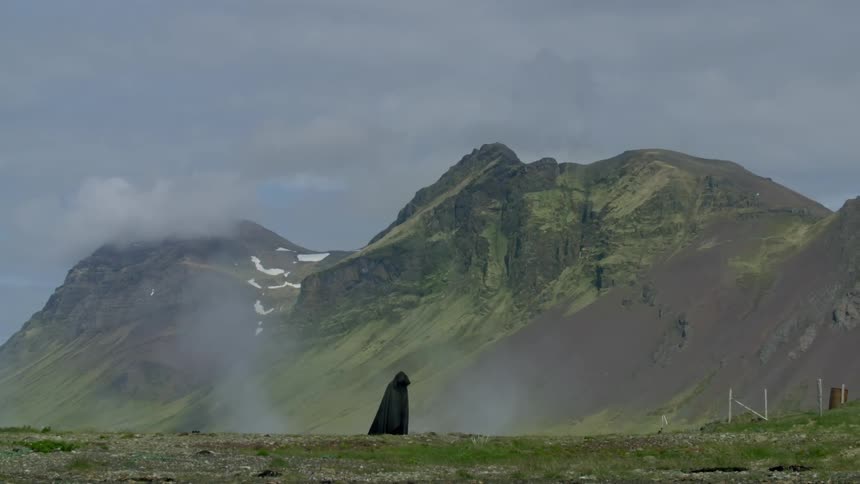 A figure dressed in a long black cape walks slowly across a field and there are mountains in the background
