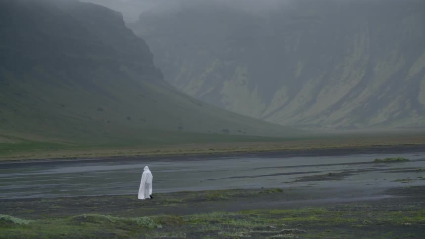 A white caped figure stands on a mountain river bank