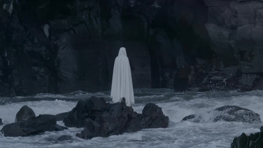 A white-cloaked figure standing on a rock formation surrounded by waves