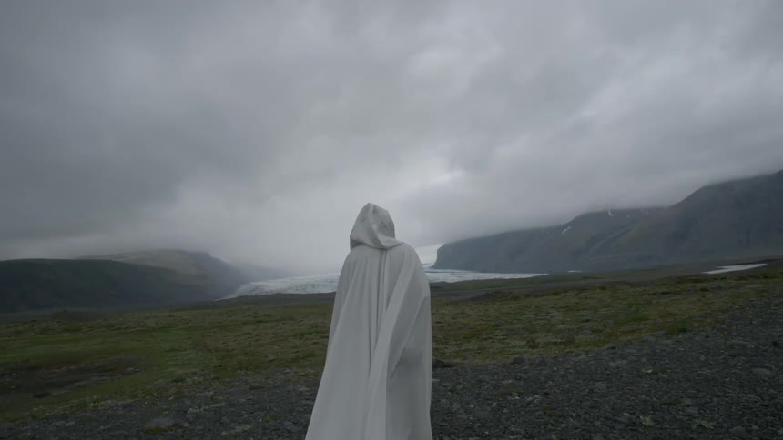 A white-cloaked figure standing in a field near mountains