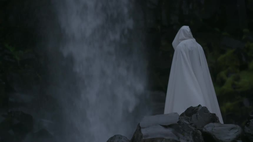 Person in white cloak standing near waterfall