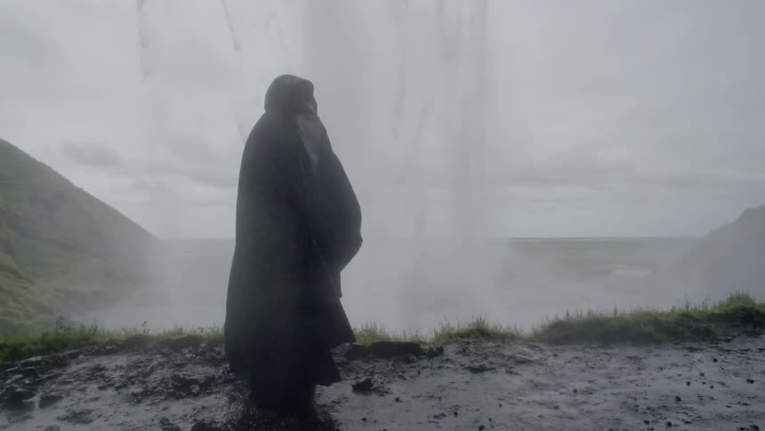 A cloaked person stands in the mist from a waterfall