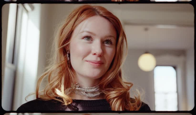 Red haired woman smiles and looks around standing near window
