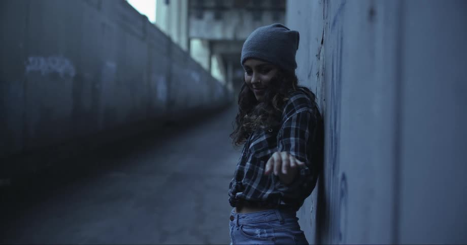 Brunette woman dances by leaning against the wall and smiles slightly in a building