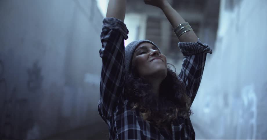 A woman stands outside a large building and throws her hands up with a large smile on her face