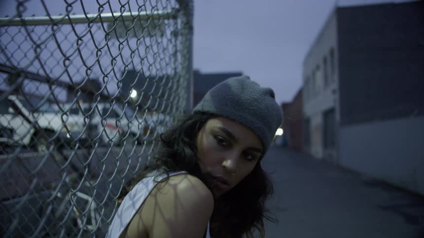 A woman leaning against a chain link fence in an alleyway in the evening