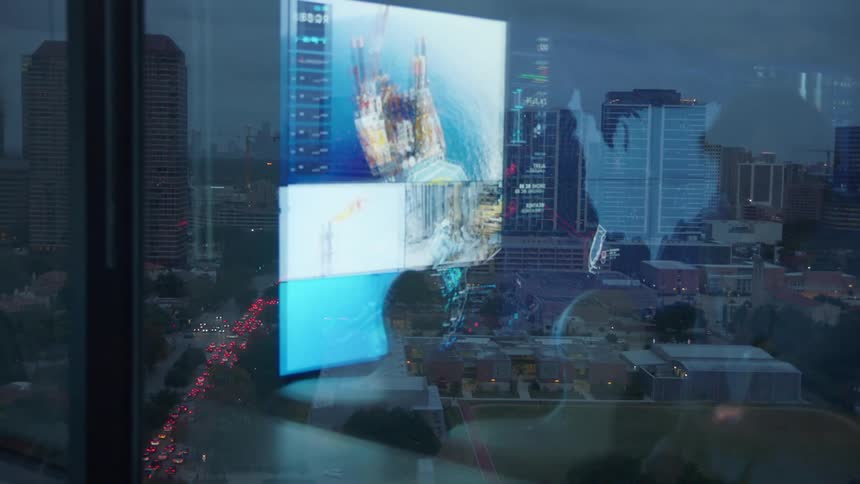 A man using his cellphone in front of the window with the city and the reflection of him and his television in it