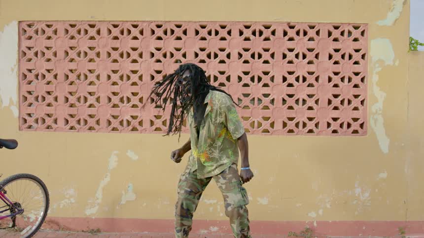 Black man with long hair performs dance moves in front of a paint chipped wall