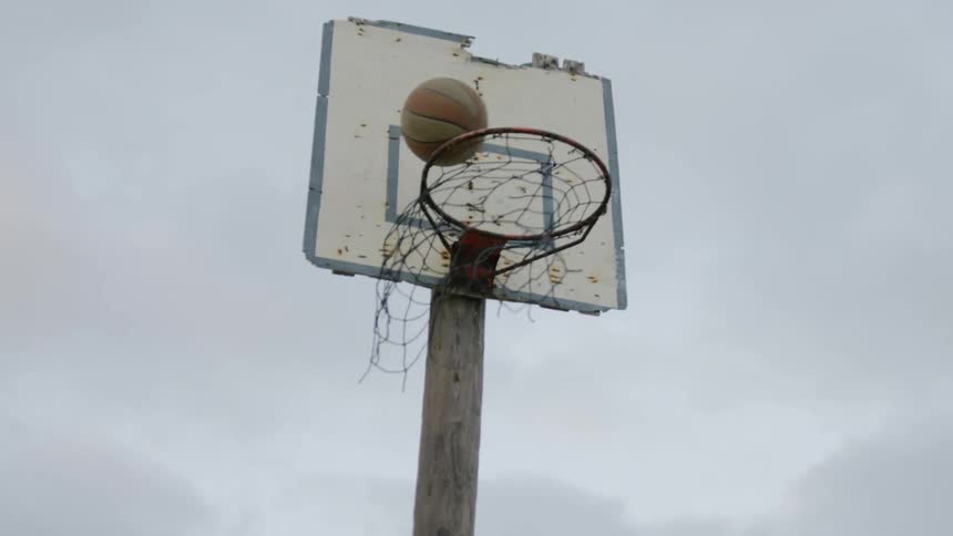 Male runs up to a worn out basketball net to thrown the ball into the hoop and the ball drops out of the bottom of the net to complete the shot