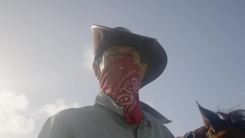 Cowboy wearing a red scarf sits on a horse and stares down at the camera