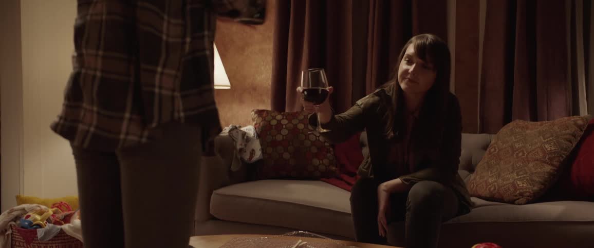 Female holding a bottle of wine stands in front of female friend who is sitting on the couch talk as she holds out a wine glass for more wine to be added