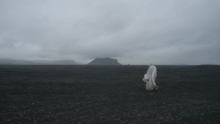 A white cloaked person walks across a barren and rocky area