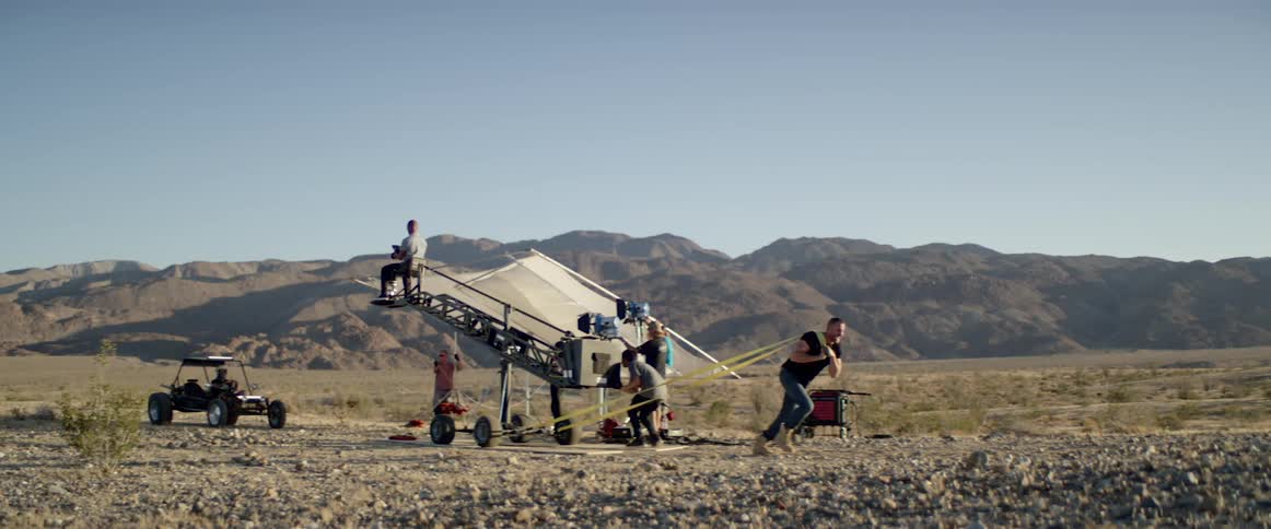 A white man pulls a large object on wheels as others film from it
