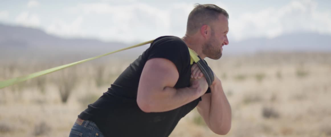 A strong white man pulls an object attached to straps across the desert