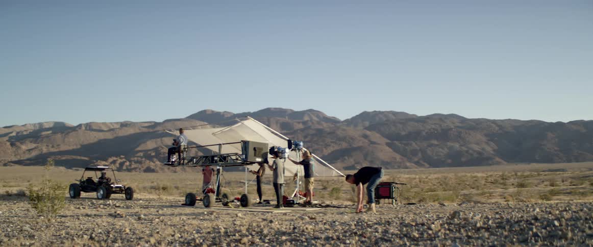 A man bends down and picks up straps while a film crew with lights and cameras looks in the direction of a parked dune buggy