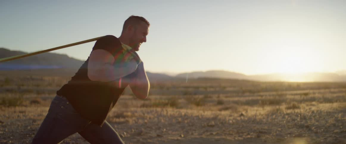 Male holding two straps over his shoulder out in the desert struggles as he tries to pull something heavy behind him as the sun is setting over the hill in the horizon