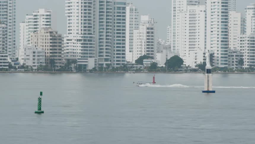 A boat cuts across the water past buoys near the city skyline
