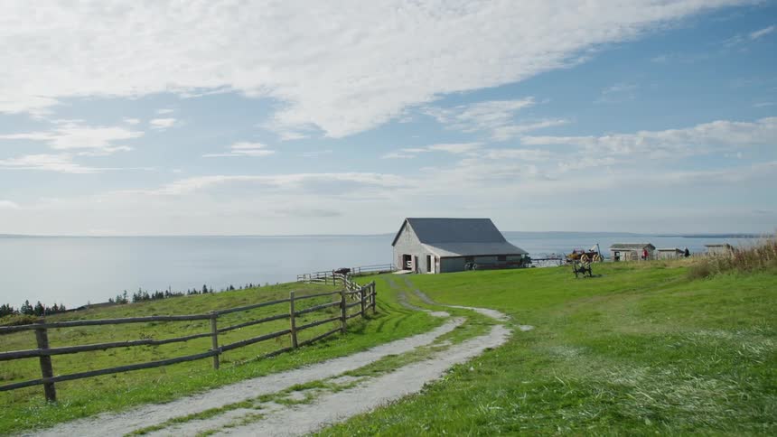 A dirt road with grass in the center leads to fenced fields and a farmhouse near the ocean