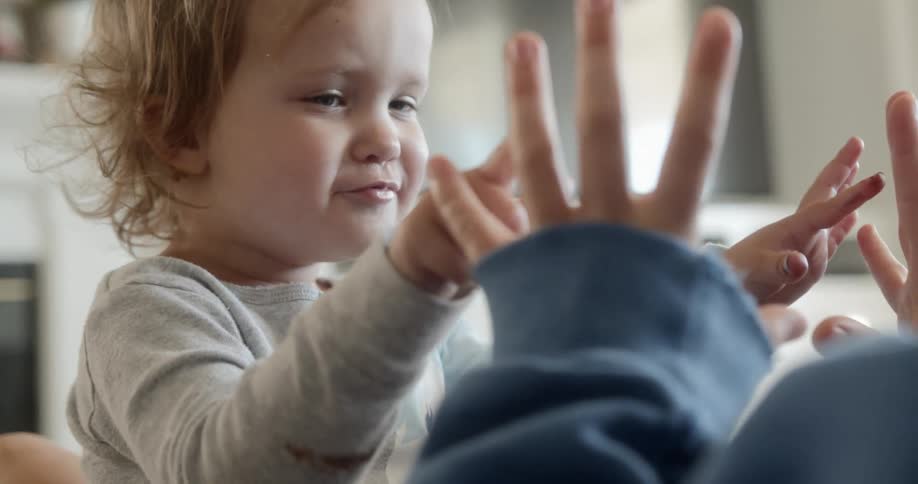 A little girl sitting on her parents lap holds up her hands and smiles as her mom does the same