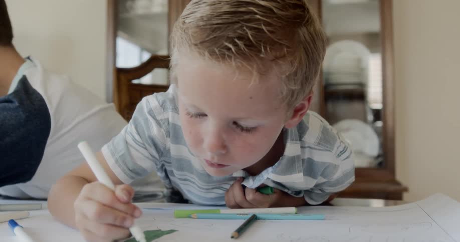 A boy sitting at a table next to another boy coloring a coloring book with a green marker