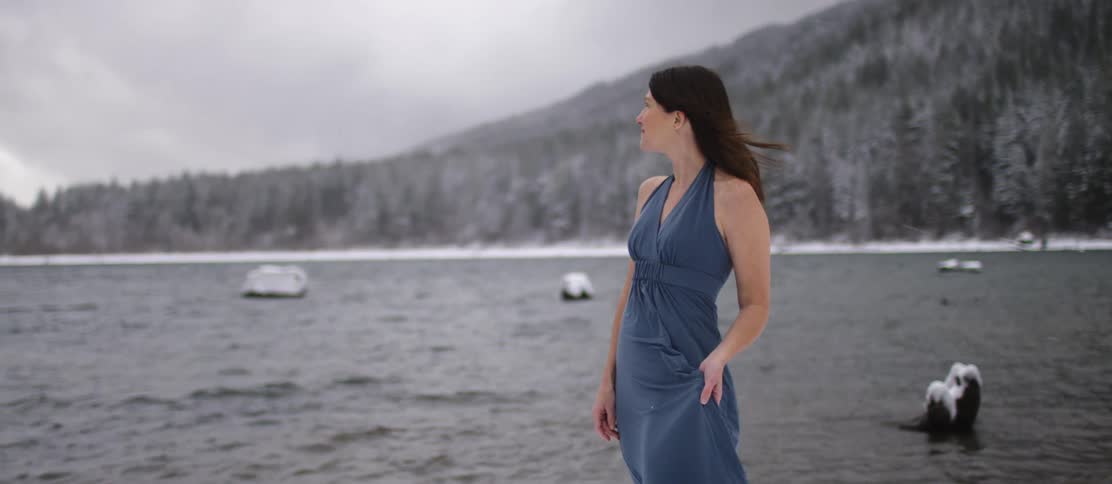 A woman poses on the shore wearing a summer dress as snow falls