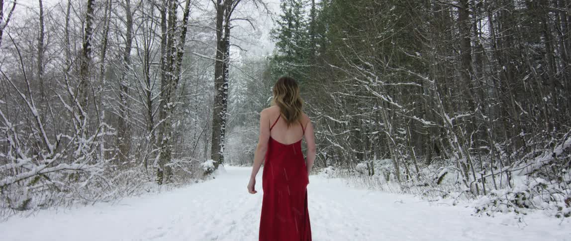 A woman in a red backless dress walking down a snow covered road as snow falls around her