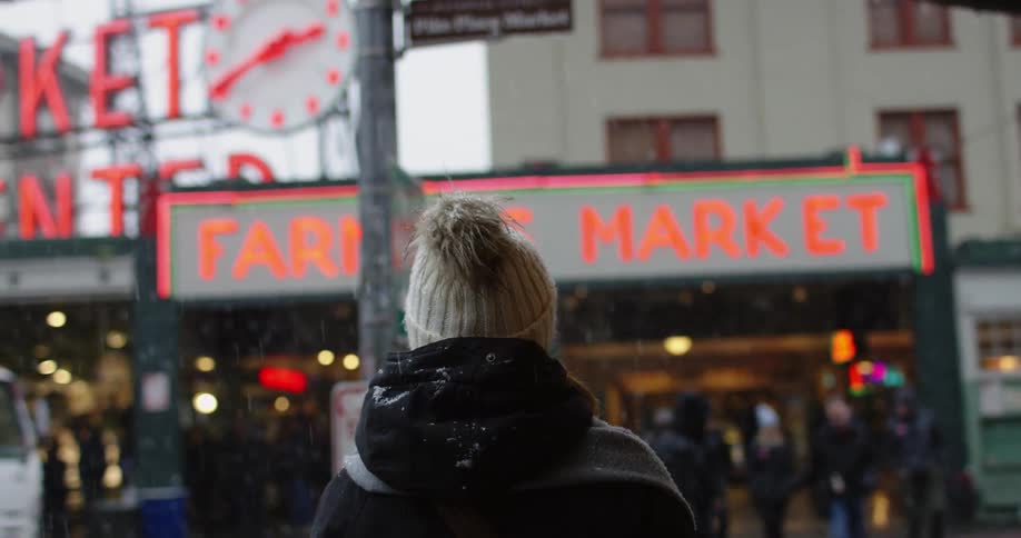 A woman in winter clothing walking across the street towards a farmers market on a snowy day