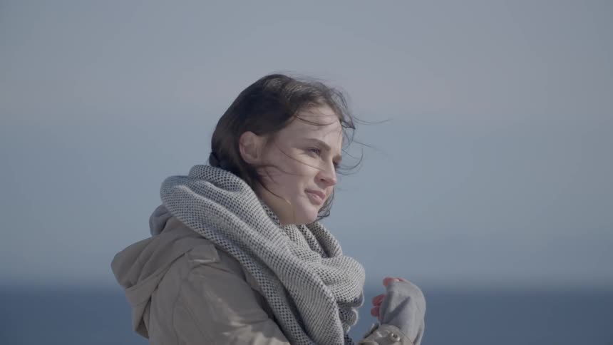 A woman wearing a heavy coat stands slightly smiling and staring out to sea