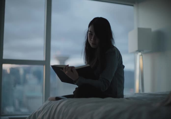 A young woman sitting on a bed looking out at the city through a large window before turning to read a book
