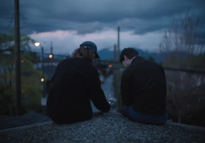 Two friends sit on the rooftop of a building and use a smartphone