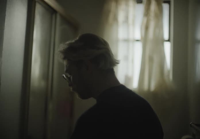 A man in a bathroom with blonde hair and mustache turns his head and stares