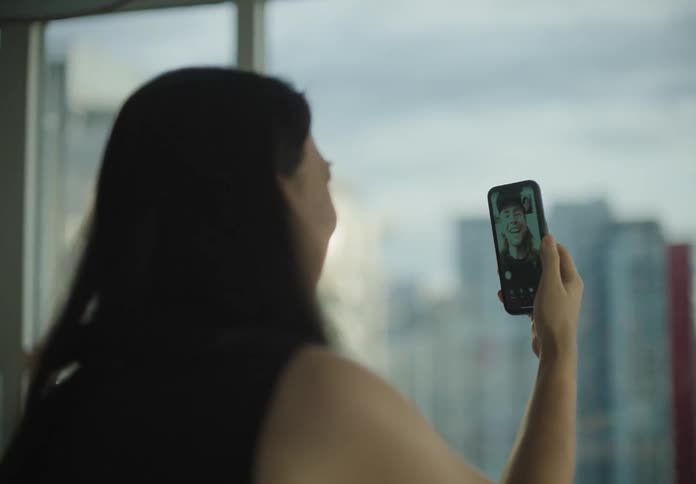 A woman picks up a cell phone and makes a video call sitting beside a large window