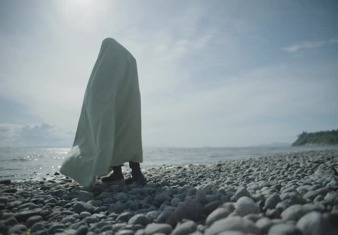 A person covered in a sheet stands on a rocky beach