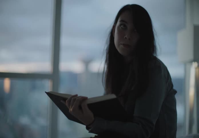 A female with long dark hair reads a book in front of a wall covered in window with a view of the space needle in the background