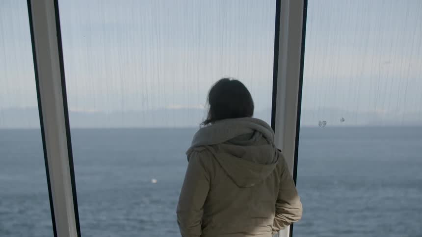 Woman stands beside large windows looking at the water